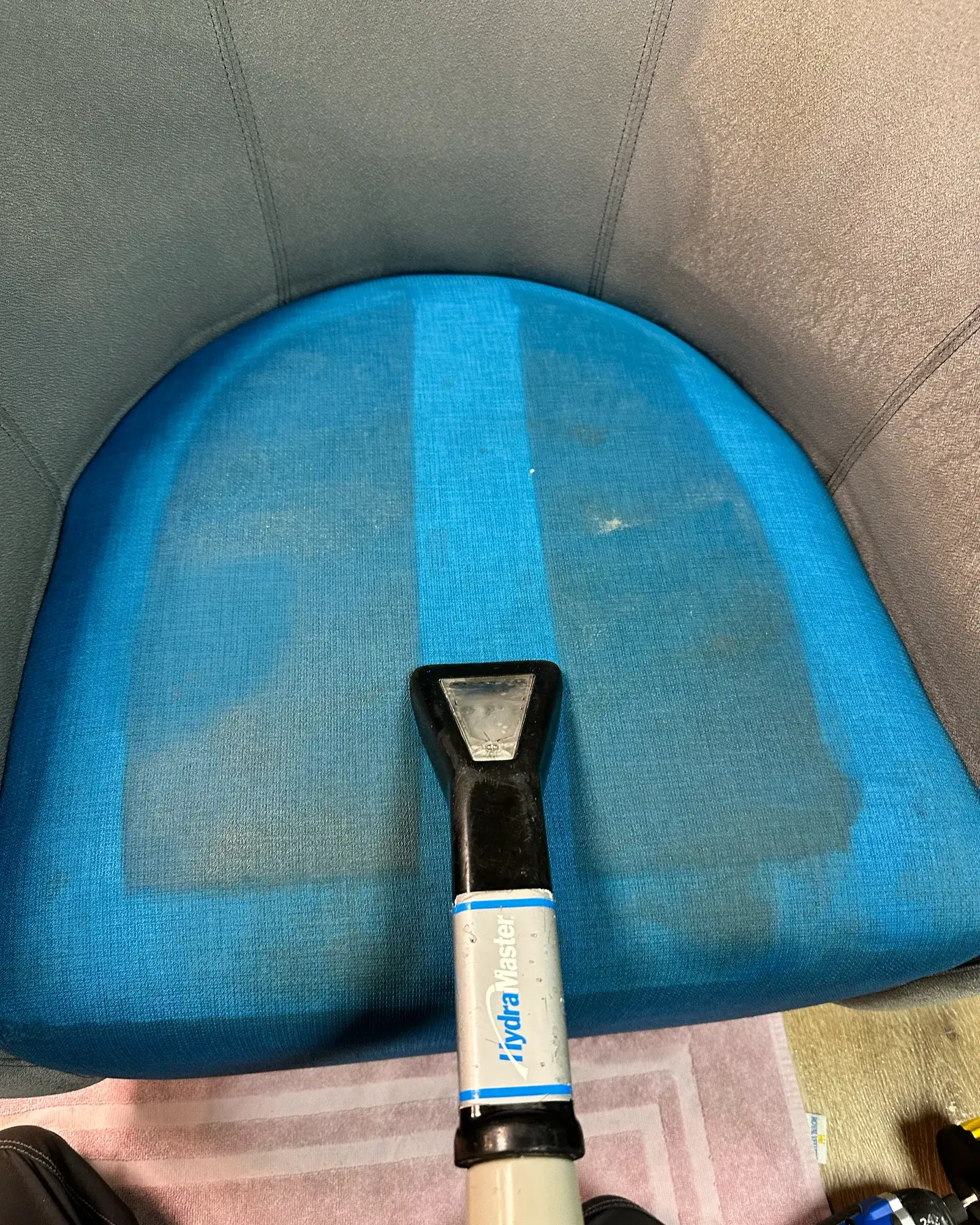 An upholstery tool part-way across the blue seat cushion of an armchair, leaving a brighter cleaned stripe beside the duller, soiled fabric.
